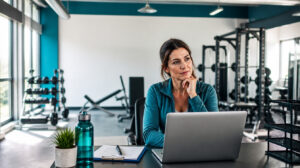 Nachdenkliche Studioleiterin sitzt mit einem Laptop an ihrem Arbeitsplatz in einem modernen Fitnessstudio.